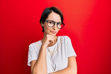 Young brunette woman with short hair wearing casual clothes and glasses smiling looking confident at the camera with crossed arms and hand on chin. thinking positive.