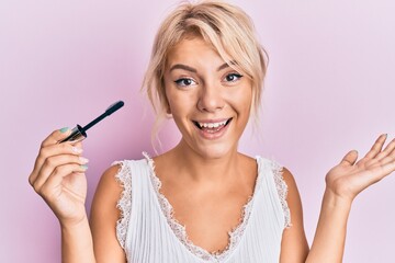 Young blonde girl holding eyelash curler celebrating achievement with happy smile and winner expression with raised hand