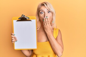 Young blonde girl holding clipboard covering mouth with hand, shocked and afraid for mistake....