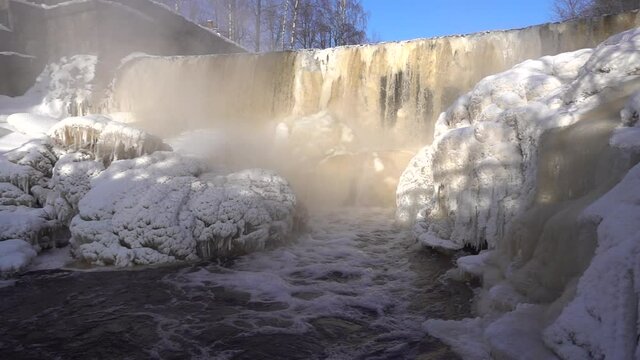 Helsinki, Finland - February 18, 2021: Water pours in slow motion over the dam in the mouth of Vantaa River at the frozen Vanhankaupunginkoski rapids (Vanhankaupunginkosken putous) on cold day.
