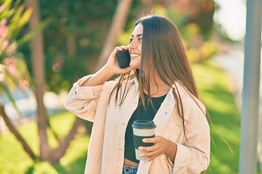 Young hispanic girl smiling happy talking on the smartphone and drinking take away coffee at the park.