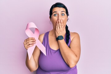 Plus size brunette woman holding pink cancer ribbon covering mouth with hand, shocked and afraid for mistake. surprised expression
