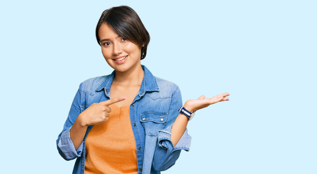 Young beautiful hispanic woman with short hair wearing casual denim jacket amazed and smiling to the camera while presenting with hand and pointing with finger.
