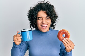 Young hispanic woman with curly hair drinking a cup of coffee and croissant winking looking at the...