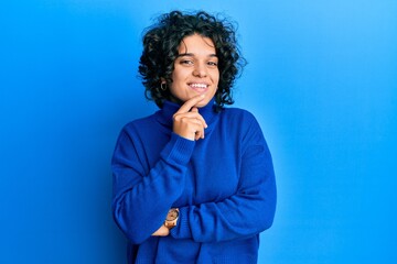 Young hispanic woman with curly hair wearing casual winter sweater smiling looking confident at the camera with crossed arms and hand on chin. thinking positive.
