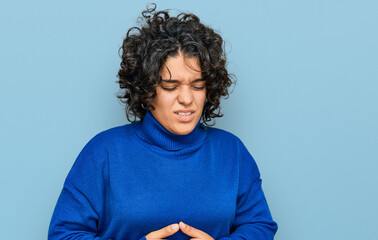 Young hispanic woman with curly hair wearing turtleneck sweater with hand on stomach because...