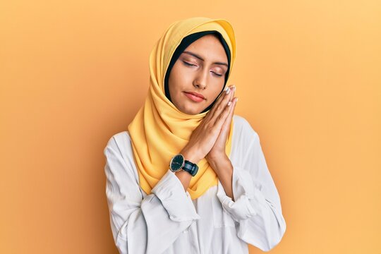 Young brunette arab woman wearing traditional islamic hijab scarf sleeping tired dreaming and posing with hands together while smiling with closed eyes.
