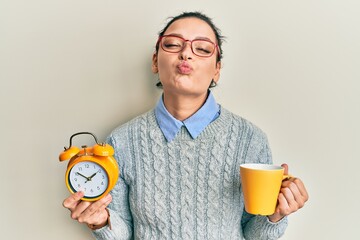 Young caucasian woman holding alarm clock drinking coffee looking at the camera blowing a kiss being lovely and sexy. love expression.