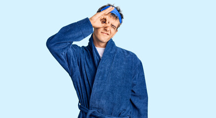 Young handsome man wearing sleep mask and bathrobe smiling happy doing ok sign with hand on eye looking through fingers