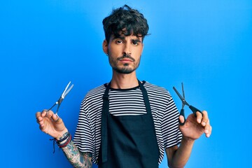 Young hispanic man wearing barber apron holding scissors relaxed with serious expression on face. simple and natural looking at the camera.