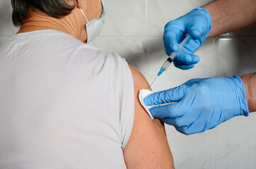 Elderly woman in medical mask receives vaccine in hospital. Blue-gloved hands inject vaccine into the woman's forearm. Concept of vaccination, coronavirus Covid19.