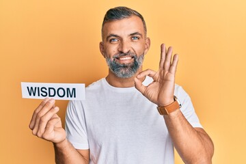 Middle age handsome man holding wisdom word on paper doing ok sign with fingers, smiling friendly gesturing excellent symbol