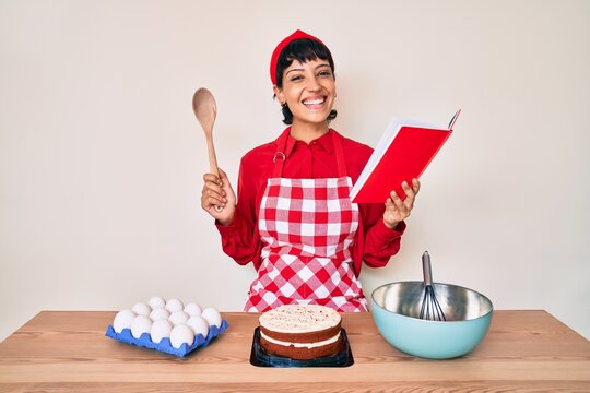 Beautiful brunettte woman cooking cake reading recipes book smiling with a happy and cool smile on face. showing teeth.