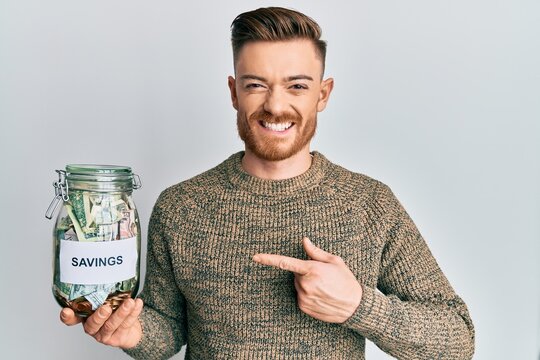 Young redhead man holding jar with savings smiling happy pointing with hand and finger