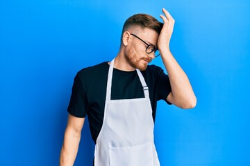 Young redhead man wearing professional apron surprised with hand on head for mistake, remember...