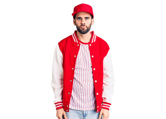 Young handsome man with beard wearing baseball jacket and cap with serious expression on face. simple and natural looking at the camera.