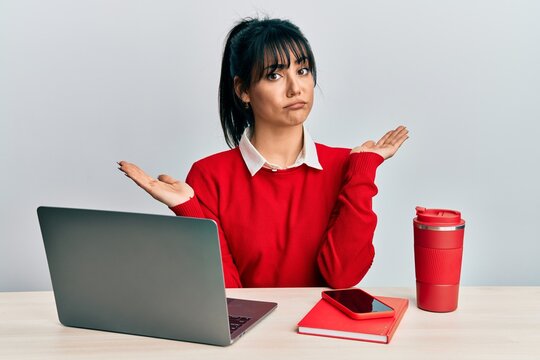 Young brunette woman with bangs working at the office with laptop clueless and confused with open arms, no idea and doubtful face.
