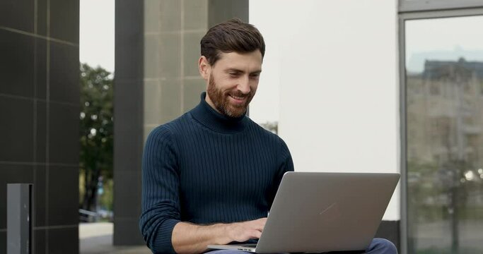 Handsome bearded man in blue sweater sitting outdoors and using portable laptop. Smiling male working on wireless computer on fresh air.