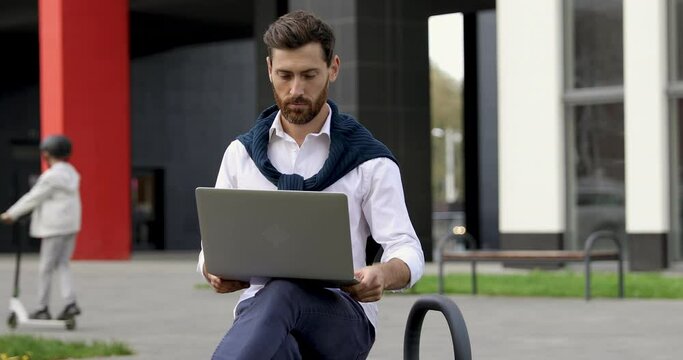 Handsome bearded man in blue sweater sitting outdoors and using portable laptop. Smiling male working on wireless computer on fresh air.