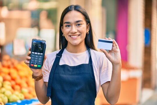 Young latin shopkeeper girl smiling happy holding credit card and dataphone at fruit store.