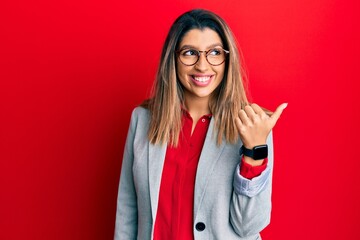 Beautiful brunette woman wearing business shirt and glasses pointing thumb up to the side smiling happy with open mouth