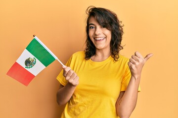 Young hispanic woman holding mexico flag pointing thumb up to the side smiling happy with open mouth