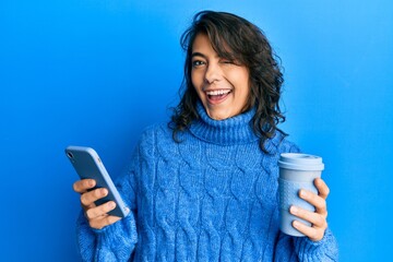 Young hispanic woman using smartphone and drinking a cup of coffee winking looking at the camera...