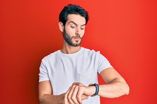 Young hispanic man wearing casual white tshirt checking the time on wrist watch, relaxed and confident