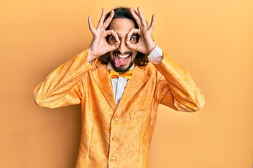 Young hispanic man wearing hipster elegant look doing ok gesture like binoculars sticking tongue out, eyes looking through fingers. crazy expression.