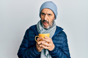 Middle age hispanic man wearing winter scarf and drinking a cup of hot coffee in shock face,...