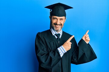 Middle age hispanic man wearing graduation cap and ceremony robe smiling and looking at the camera pointing with two hands and fingers to the side.