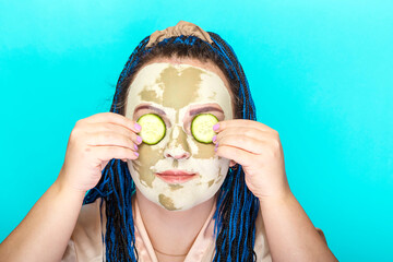 Woman with blue afro braids face in a mask made of green clay with cucumber circles on her eyes on a blue background.