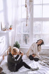 Two young girls are photographed in a white room on the bed near the window