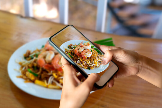 Hand holding mobile phone and take a photo Fried rice with spicy shrimp on blurred wooden table  background with sunlight.