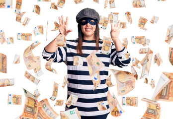 Young beautiful brunette woman wearing burglar mask showing and pointing up with fingers number...
