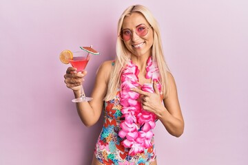 Young blonde woman wearing swimsuit and hawaiian lei drinking cocktail smiling happy pointing with hand and finger