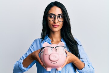 Beautiful hispanic woman holding piggy bank with glasses smiling looking to the side and staring away thinking.
