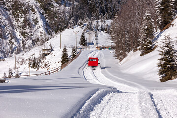 Red snowcat is going towards the ski resort. Smoking snow vehicle cleared the snow on the road already and left a clean road behind. The valley in the mountains is under heavy snow.