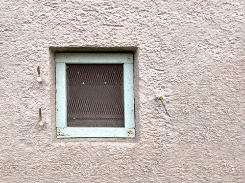 Kleines quadratisches Fenster mit wei&szlig;em Holzrahmen mitdunkelbraunen Holz verschlossen, in  hellrosa Hausfassade . Scharniere f&uuml;r Fensterladen sind an der Hauswand befestigt, jedoch ohne Laden