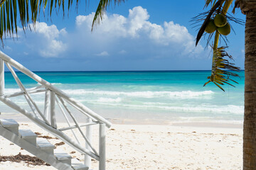 A white wooden staircase on a white wooden staircase in a Caribbean beach in Mexico with palm trees and coconuts. In the background the Ocean and partly cloudy sky. The ideal place for vacation.