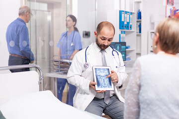 Obraz premium Healthcare specialist explaining senior woman diagnosis of braing trauma on tablet pc. Medical therapist showing and talking about x-ray with patient during consultation.