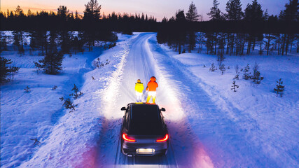 Aerial back view of romantic couple of tourists standing near car while headlights lighting them way on in winter darkness, bird's eye view of vehicle in snowy north lands. Wanderlust in road trip