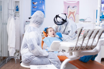 Dental specialist in the course of little girl oral hygine consultation wearing ppe suit as safety precaution because of covid19. Dentist in coronavirus suit using curved mirror during teeth