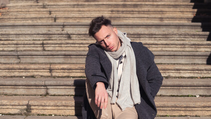 Handsome young man outdoor wearing coat, scarf and shirt sitting on stone stairs outside, looking at camera