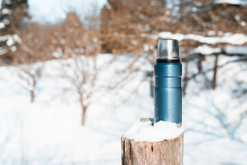 Blue vacuum travel flask. Thermos covered with snow, tree stump, winter forest on a sunny day.