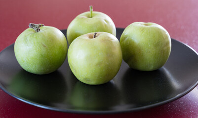 A few green fresh apples lie on a black platter on the red table.