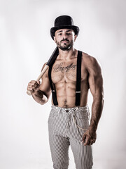 Handsome shirtless muscular man standing on white background, wearing bowler hat and suspenders on naked torso, holding baseball bat