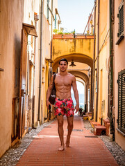 Handsome muscular shirtless man walking on old alley in Italian town in summer wearing swimming shorts. Vertical outdoors shot