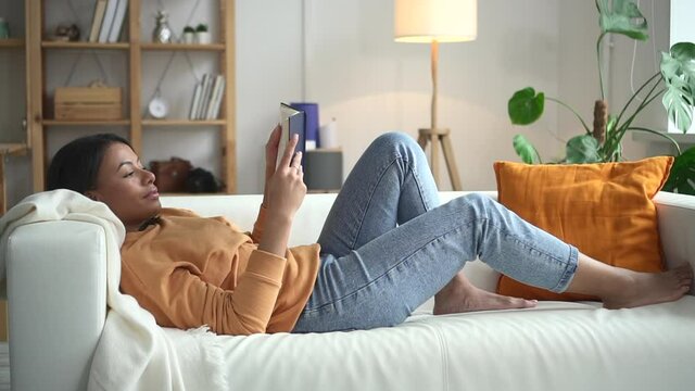 African woman is reading book on sofa in bright room Spbd. Black-skin female model is passionate about adventure, travel and fashion literature. Reader took an interesting press from library for