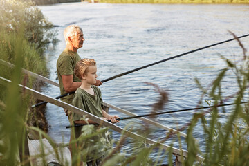 European grey haired mature father with son outdoors fishing by lake or river, standing near water with fishing rods in hands, dress casually, enjoying hobby and nature.